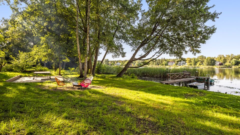 Grüne Uferwiese an einem ruhigen See mit hohen Laubbäumen und Sitzbereich im Schatten. Rechts führt ein kleiner Holzsteg ins Wasser, daneben liegt ein Boot am Schilfrand; gegenüber sind einzelne Häuser am Ufer zu sehen.