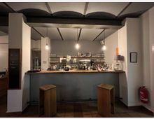 Eine kleine, minimalistisch gestaltete Bar mit schlichten weißen Wänden, hölzerner Theke und zwei Holzhockern. Im Hintergrund sind Regale mit Gläsern und Flaschen sowie eine Kaffeemaschine sichtbar, beleuchtet von hängenden Glühbirnen.