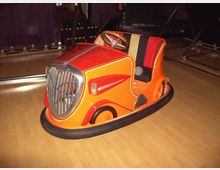 Ein orangefarbener Autoscooter im Retro-Design mit verchromtem Kühlergrill und rot-schwarzen Sitzen steht auf einem Holzboden in einer Halle. Im Hintergrund sind Bühnenelemente und metallische Konstruktionen zu sehen.