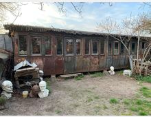 Verrosteter, stillgelegter Waggon mit mehreren Fenstern in einem verwilderten Außenbereich, umgeben von kargen Bäumen und Gebüsch; im Vordergrund liegen mehrere Skulpturen von Köpfen sowie gestapelte Holz- und Metallteile.