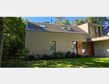 Modernes Wohnhaus mit flachem, schrägem Dach und großen Fenstern, umgeben von einem Garten mit Rasen, Büschen und Bäumen. Das Gebäude hat eine beige Fassade und hölzerne Türelemente, mit einem ruhigen Waldumfeld im Hintergrund.