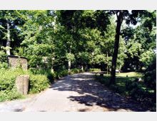 Ein schattiger Parkweg aus Kopfsteinpflaster, umgeben von hohen, dicht belaubten Bäumen und Grünflächen, mit einem alten, niedrigen Ziegelbau links im Hintergrund.