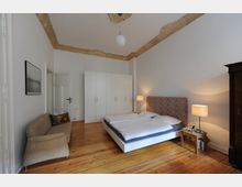 Schlafzimmer mit Holzboden, einem weißen Doppelbett mit gemustertem Kopfteil, zwei Nachttischen mit Lampen, einem beige Sofa, einem weißen Kleiderschrank und einer dekorativen Stuckverzierung an der Decke. Die Wände sind weiß gestrichen, und das Zimmer wirkt geräumig.