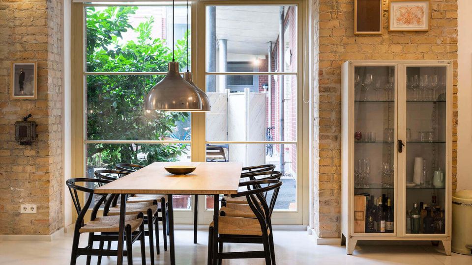 Interior dining area with exposed brick walls, a wooden table and black chairs beneath a hanging metal pendant light. Large floor-to-ceiling glass doors and windows open to a small courtyard with greenery, and a white glass-front cabinet displays glassware and bottles.
