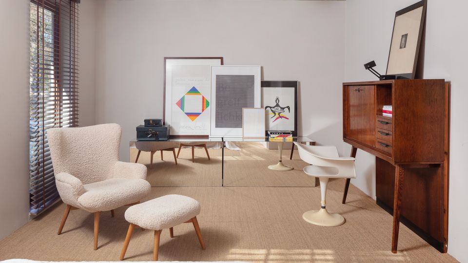 Minimalist bedroom or sitting area with a concrete ceiling, white walls, and woven carpet, lit by a window with dark blinds. Furnishings include a cream lounge chair with ottoman, two white pedestal chairs, a wood cabinet, and several framed artworks leaning against the back wall.