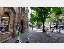 Straßenszene in einer Stadt mit einem historischen Backsteingebäude auf der linken Seite, verziert mit Ornamenten und Graffiti. Im Hintergrund sind Bäume, geparkte Fahrräder, Elektroroller, Geschäfte und eine erhöhte Bahntrasse sichtbar.