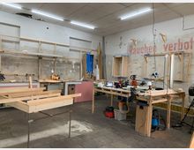 Ein Handwerksraum mit Arbeitsflächen aus Holz, Holzrahmen, Werkzeugen und Maschinen. An der Wand ist der Schriftzug „Rauchen verboten“ zu sehen, daneben Regale und provisorische Konstruktionen in einer Werkstattumgebung.