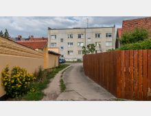 Ein schmaler Weg führt zwischen einer gelben Mauer mit Sonnenblumen und einem hohen Holzzaun zu einem mehrstöckigen Wohngebäude aus grauem Beton im Hintergrund. Im Hof sind geparkte Autos und etwas Grün zu sehen, daneben stehen kleine Bäume.