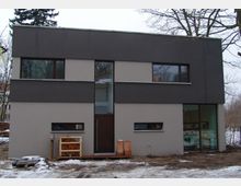 Modernes zweistöckiges Wohnhaus mit flachem Dach, grauer und cremefarbener Fassade sowie großen, rechteckigen Fenstern. Im Vordergrund ist ein schneebedeckter Boden und ein Stapel Brennholz unter einem kleinen Regaldach zu sehen.