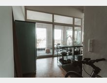 Ansicht eines Wohnungsbereichs mit Blick auf einen Balkon. Im Vordergrund ein Raum mit Holzfußboden, einem grünen Schrank, einem schwarzen Metallregal mit Küchenutensilien und einem Fahrradlenker; im Hintergrund große Glas-Schiebetüren, die zu einem gefliesten Balkon mit Pflanzen und Möbeln führen.