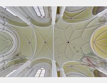 Decke eines historischen Kirchengebäudes mit grünlich-gelben Gewölben, verzierten weißen Rippen und Spuren von Verwitterung. Große, bogenförmige Fenster mit vergittertem Glas lassen Licht in den Innenraum.