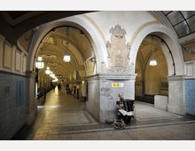 Ein historischer U-Bahnhof mit gewölbten Decken, dekorativen Steinornamenten und hängenden Leuchten. Im Vordergrund sitzt ein Straßenmusiker mit einem Akkordeon neben einer Säule, die mit Reliefs und Graffiti verziert ist.