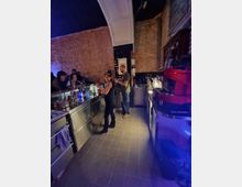 Eine Bar mit freiliegendem Backstein und gewölbter Decke, in der zwei Personen hinter der Theke arbeiten; Edelstahl-Arbeitsflächen, Flaschen und eine rote Espressomaschine sind sichtbar. Im Hintergrund sind Gäste und blaues Licht zu erkennen.