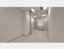 Ein leerer, rechteckiger Raum mit weißen Fliesenboden und weiß gestrichenen Wänden. Eine große Fensterfront mit vertikalen Jalousien lässt natürliches Licht herein, und an der Wand darunter befindet sich ein Heizkörper. Deckenplatten und Deckenbeleuchtung sind sichtbar.