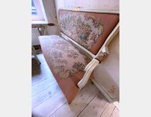 Antikes Sofa mit einer rosafarbenen Polsterung, verziert mit floralen Stickereien und einer Darstellung von Personen in einem ländlichen Szene-Motiv; weißes, geschnitztes Holzgestell mit goldenen Details, auf hellem Holzboden in einem Raum mit schlicht verputzten Wänden.