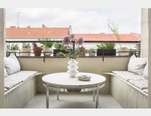 Kleiner Balkon mit seitlichen Sitzbänken, hellen Polsterkissen und einem runden Tisch in der Mitte. Auf dem Tisch steht eine weiße Vase mit Blumen, umgeben von Blumentöpfen auf der Balkonbrüstung; im Hintergrund rote Ziegeldächer und blauer Himmel.