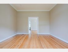 Leerer, heller Wohnraum mit hohen Decken, weißer Stuckleiste und breiten Dielen-Holzboden. In der Mitte führt eine Türöffnung in einen weiteren Raum mit ähnlichem Boden und einem Fenster im Hintergrund.