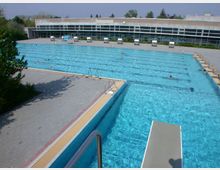 Großes Freibad mit einem rechteckigen Schwimmbecken, mehreren Schwimmbahnen und einem Sprungbrett im Vordergrund. Im Hintergrund ein modernes Gebäude mit großen Fenstern und einer umliegenden Terrasse mit Sonnenliegen.