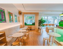 Bright, modern café interior with light wood floors, round white tables, and bench seating along a wall of framed posters. Large front windows include a built-in window seat with cushions, and a green espresso counter sits near the entrance with additional seating and indoor plants.