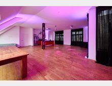 Weitläufiger Innenraum mit Holzboden, einer offenen Küchenzeile mit roten Elementen sowie schwarzen Vorhängen vor großen Fenstern. Die Beleuchtung ist in violett und rosa gehalten, im Vordergrund steht ein großer Holztisch.