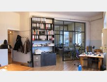 Ein Büro mit Holzboden, einer Glastrennwand, die den Raum teilt, sowie offenen Regalen mit Ordnern und Büchern an der Wand. Im Vordergrund stehen Schreibtische mit Arbeitsmaterialien, und im Hintergrund sind Bürostühle und Pflanzen sichtbar.