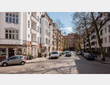 Straßenszene in einer Wohngegend mit gepflasterter Straße, mehrstöckigen Gebäuden mit weißer und roter Fassade sowie Balkonen. Links sind Geschäfte und Cafés zu sehen, rechts und entlang der Straße geparkte Autos, im Hintergrund ein größeres Backsteingebäude.