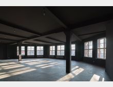 Leerer, dunkler Raum mit großen Fenstern, die den Raum mit natürlichem Licht durchfluten. Deckenstützen und ein Betonboden dominieren die minimalistische Architektur.