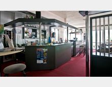 Barbereich in einem Innenraum mit rotem Teppichboden, dunkelgrauer Tresenfront und mehreren Hockern. Im Hintergrund sind Regale mit Flaschen, Fenster mit Tageslicht sowie einige Tische und Stühle sichtbar.