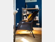 Ein modern eingerichteter Raum mit blau gestrichener Wand, einem schwarzen Netzsessel im Vordergrund und einem Regal mit Büchern, Dekofiguren sowie einer Tischleuchte an der Wand. Der Boden besteht aus hellem Holzparkett.