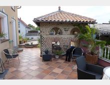 Geräumige Terrasse mit terrakottafarbenen Fliesen und einem kleinen Pavillon aus Naturstein mit Rundbögen und Ziegeldach. Der Bereich ist mit Gartenmöbeln, Pflanzen in Töpfen und dekorativen Elementen gestaltet.
