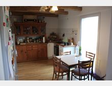 Küche mit rustikalem Holzschrank, der als Stauraum für Geschirr und Lebensmittel dient, und einer weißen Küchenzeile mit eingebautem Herd. Im Vordergrund steht ein Esstisch mit vier Holzstühlen, dahinter eine große Glastür, die viel Tageslicht hereinlässt.