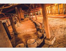 Innenansicht einer historischen Fabrikanlage mit einer Backsteinbodenfläche, massiven Holzträgern und mehreren Metallkesseln, die entlang eines gewölbten Pfades aufgereiht sind. Im Hintergrund sind Backsteinwände, Beleuchtung und weitere industrielle Elemente sichtbar.