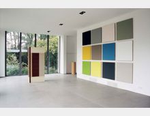 Ein moderner Ausstellungsraum mit weißen Wänden und großen bodentiefen Fenstern, die den Blick auf Bäume und Grünflächen freigeben. An der rechten Wand hängt eine quadratische Anordnung farbiger Leinwände, während in der Mitte des Raums ein schmaler rechteckiger Holz- und Metallaufsteller steht.