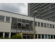 Flaches Gebäude mit großen Fenstern, auf denen der Schriftzug „Sommerfest 2. Juni 10–14 Uhr“ angebracht ist, im Vordergrund ein Eingang mit Glasfront und Pflanzen im unteren Bereich; im Hintergrund ein graues Hochhaus und eine Straßenlaterne.