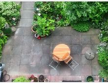 Vogelperspektive eines bepflanzten Gartens mit Steinplatten und einem kleinen Sitzbereich aus einem runden Holztisch und zwei Klappstühlen. Rundherum stehen zahlreiche Pflanzen in Töpfen und Beeten, die Fläche ist von grüner Vegetation umgeben.