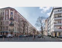 Straßenszene mit mehrstöckigen Wohngebäuden im Altbau-Stil auf der linken Seite und modernen Gebäuden mit Balkonen auf der rechten Seite. Geparkte Autos, ein Fußgängerüberweg und Bäume säumen die Straße, während ein Geschäft im Erdgeschoss sichtbar ist.