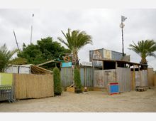 Eine Strandbar mit improvisierten Gebäuden aus Holz und Containern, umgeben von sandigem Boden, Bambuszäunen und Palmen. Im Hintergrund sind grüne Bäume und eine leicht bewölkte Himmel zu sehen.