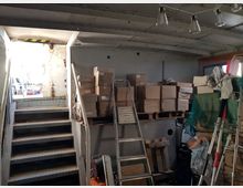 Ein Metallraum mit einer Treppe, die zu einer hell erleuchteten Tür führt. Im Raum befinden sich gestapelte Kartons auf Paletten, eine Leiter und verschiedene Werkzeuge sowie Gegenstände wie Netze und Kabel.