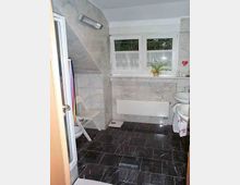 Badezimmer mit dunklem Marmorboden und hellen Wandfliesen, einem Fenster mit Gardinen und einer kleinen Pflanze auf der Fensterbank. Im Raum befinden sich eine Dusche, ein Waschbecken, ein Heizkörper und ein Klappstuhl.