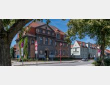 Straßenansicht mit einem historischen zweistöckigen Gebäude mit rotem Satteldach und einer Fassade in Grau- und Beigetönen, genutzt als Polizeigebäude. Im Vordergrund sind Verkehrszeichen, Bäume und Fußwege zu sehen, rechts daneben weitere Wohnhäuser.