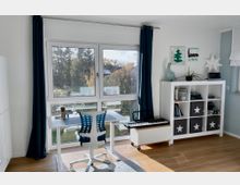 Heller Raum mit großer Fensterfront und dunklen Vorhängen, davor ein höhenverstellbarer Schreibtisch mit Bürostuhl. Rechts stehen ein weißes Regal mit Aufbewahrungsboxen und Dekoration sowie ein niedriges Möbel mit Keyboard; Holzboden und Blick ins Grüne.