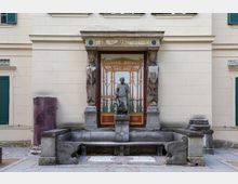 Steinbrunnen mit einer zentralen Skulptur eines Mannes in antiker Pose, flankiert von zwei Karyatiden-Säulen, vor einer aufwendig dekorierten Wand mit antikem Fresko. Die Fassade ist hell und strukturiert mit grünen Fensterläden.