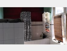 Badezimmer mit einer weißen, gefliesten Badewanne, einer schwarz-weiß gemusterten Duschvorhangstange, offenen Regalfächern mit Hygieneartikeln sowie einem Fenster mit braunen Jalousien. Die Wand ist teilweise rot und grün gestrichen.