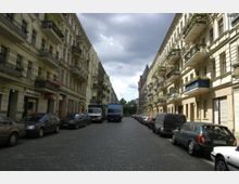 Eine gepflasterte Straßenansicht in einer Stadt mit historischen Wohngebäuden auf beiden Seiten, die mehrere Stockwerke und Balkone mit Blumenschmuck aufweisen. Am Straßenrand sind zahlreiche parkende Autos und Lieferwagen zu sehen, im Hintergrund sind Bäume und ein Turm erkennbar.