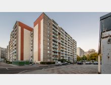 Wohnblöcke in einem urbanen Gebiet mit mehreren Stockwerken und zahlreichen Balkonen. Im Vordergrund ist ein gepflasterter Parkplatz mit geparkten Autos sowie ein Weg zu sehen; im Hintergrund stehen weitere ähnliche Gebäude und einige Bäume.