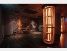 Ein stilvoll beleuchteter Innenraum mit dunklem Holzboden und einer dekorativen Wand, die mit einem kunstvollen Waldmotiv mit Tieren verziert ist. Im Vordergrund stehen moderne, säulenförmige Lichtinstallationen mit warmem, gelb-orangefarbenem Licht.