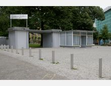 Flacher Pavillonbau mit großen Fenstern und einem Flachdach, umgeben von einem gepflasterten Platz und Bäumen im Hintergrund; auf dem Dach des Gebäudes ein Schild mit der Aufschrift "Grüner Salon".