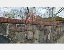 Niedrige Steinmauer aus großen, unregelmäßigen Feldsteinen mit roter Ziegelabdeckung entlang eines gepflasterten Weges. Dahinter stehen blattlose Bäume und ein Backsteingebäude mit moosigem Ziegeldach.