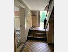 Ein schmaler Hausflur mit gelb-schwarz kariertem Fliesenboden und einem kurzen Treppenabsatz, der zu einer Glastür mit Blick auf einen Garten mit Bäumen führt. Links befindet sich eine Türöffnung zu einem weiteren Raum, und rechts sind Wandfliesen sowie eine Treppe sichtbar.