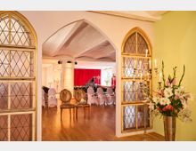 Blick durch einen Rundbogen in einen festlichen Veranstaltungssaal mit Holzboden, Stuhlreihen und einem Flügel vor roten Bühnenvorhängen. Links und rechts rahmen bleiverglaste Bogenfenster den Eingang; daneben stehen ein mehrarmiger Kerzenleuchter und eine große Blumenvase.